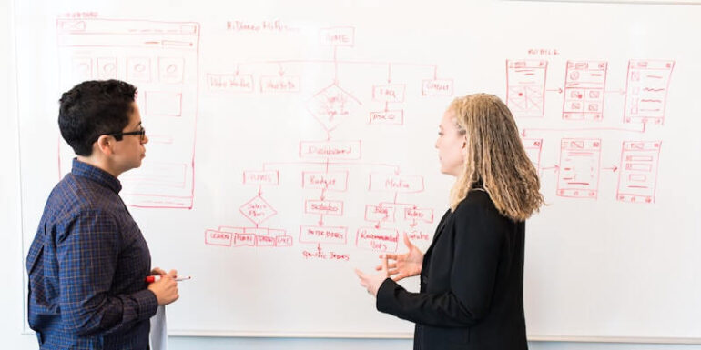 Two people talking in front of a whiteboard with a UI diagram on it.