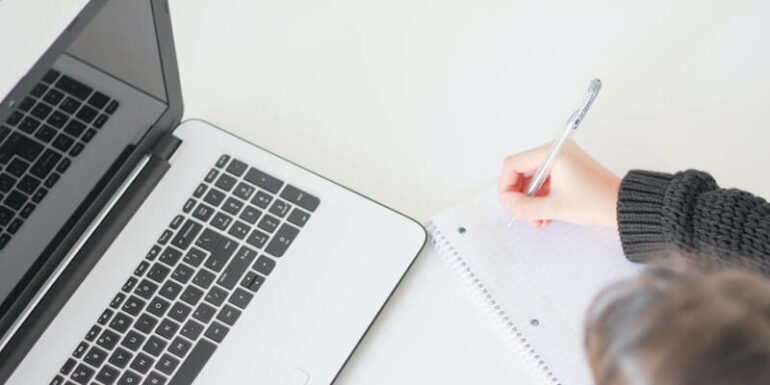 A person looking at a laptop and writing in a notebook.